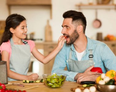 a cheerful young girl gives slice of fresh tomato to her delighted father during fun and interactive healthy cooking session