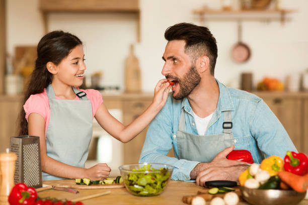 a cheerful young girl gives slice of fresh tomato to her delighted father during fun and interactive healthy cooking session