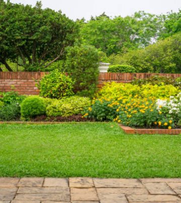 Backyard English cottage garden, colorful flowering plant and green grass lawn, brown pavement and orange brick wall, evergreen trees on background, in good care maintenance landscaping in park