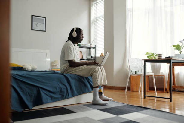 Young man wearing headphones sitting on bed and using laptop in minimalist bedroom with sunlight streaming through window, surrounded by simple furniture