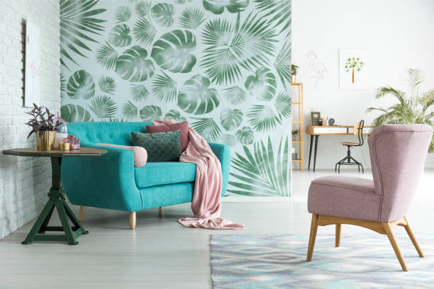 Pastel pink armchair standing in front of a blue couch with pink blanket in botanic interior of open living room